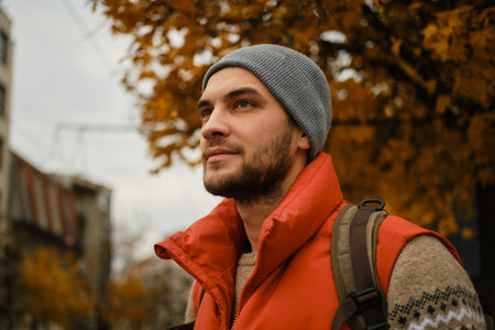 Portrait of a young man in a red vest and gray beanie, standing outdoors with autumn trees in the backgroundの写真素材