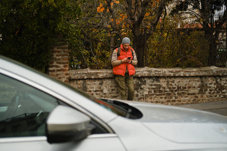 Man in casual autumn outfit leans against a brick wall, looking at his phone on a street with cars...