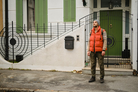 Man in red vest standing in front of white house with green shutters and gate. Autumn scene