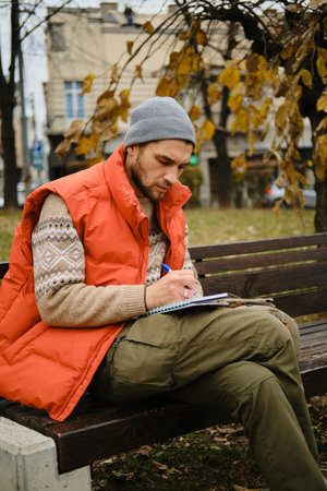 A man in a gray beanie and red vest writes notes in a notebook while sitting on a bench with his...