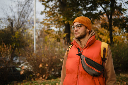 Man with glasses in autumn outfit walks down a leaf-covered park path, wearing a red vest and orange...