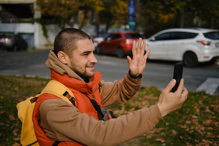 Man waving to smartphone during online video chat in autumn park. Travel blogger concept