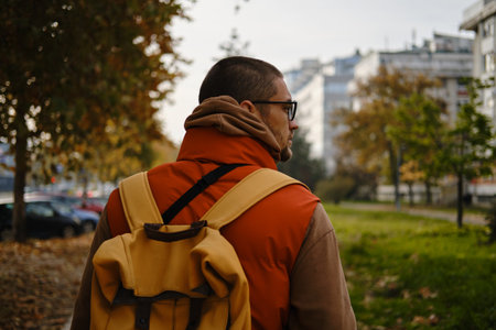 Rear view of man with yellow backpack walking in autumn park with buildings behind