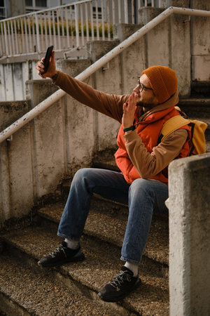 Man sitting on stairs taking selfie with smartphone, wearing orange vest and beanie. Travel blogger...