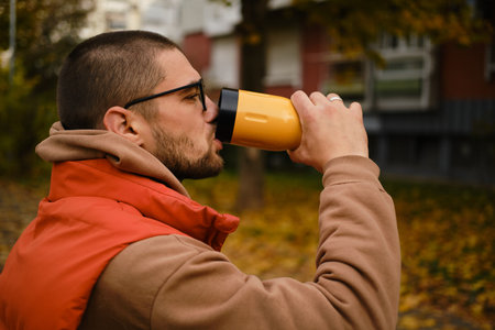 Young man drinking from travel mug while sitting on a bench in Belgrade park. Drink hot take away...