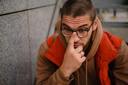 Close-up of man adjusting glasses while sitting near wall, casual autumn street style