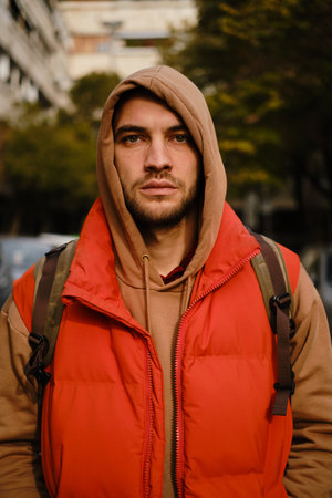 Close-up urban portrait of serious man in hood, red vest and glasses, standing on street with parked...