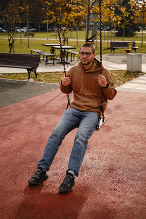 Young man in hoodie sitting on swing in park, relaxed lifestyle portrait