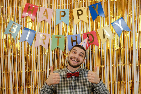 Cheerful man in party hat and red bow tie smiles with thumbs up in front of Happy Birthday banner....