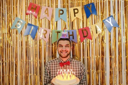 Smiling man with birthday cake and candles, party banner and golden festive backdrop