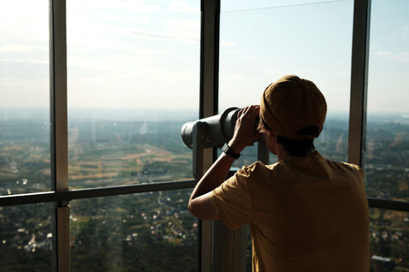 Young woman in yellow cap using a public binocular viewer at the top of Avala Tower overlooking the green hillsの写真素材