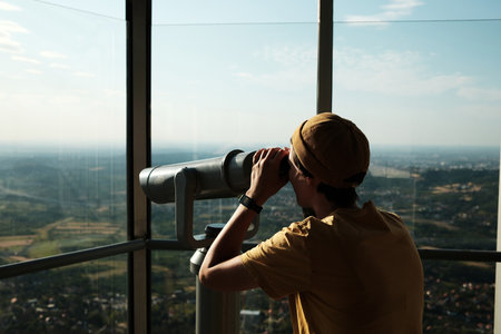 Young woman in yellow cap using a public binocular viewer at the top of Avala Tower overlooking the green hills of Belgradeの写真素材