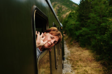 Mature woman waving out of a moving vintage train in Mokra Gora, surrounded by lush green forest in...