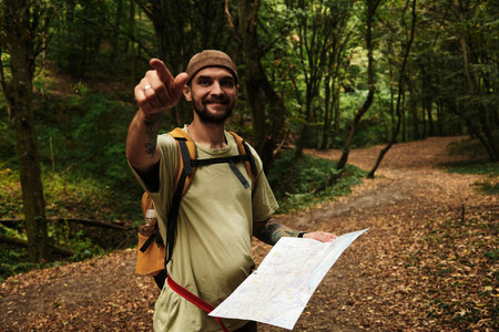 Smiling man with backpack holding map and pointing direction in Fruska Gora forest, Serbia. Front view portrait of touristの写真素材
