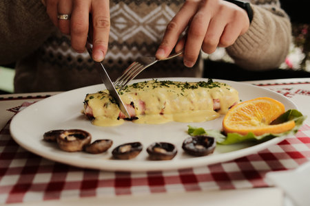 Close-up of hands cutting grilled meat dish topped with creamy cheese sauce and garnished with herbs, served with mushrooms and orange sliceの写真素材