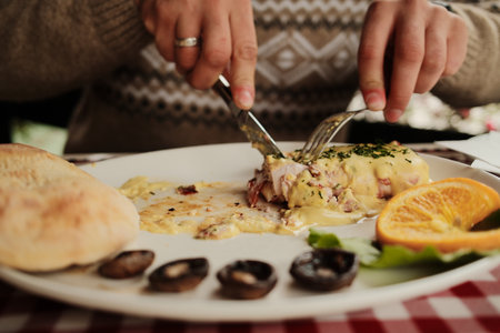 Close-up of hands cutting grilled meat dish topped with creamy cheese sauce and garnished with...