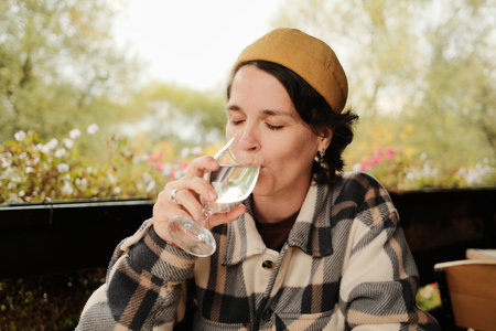 Woman smiling while holding and drinking a glass of water at an outdoor cafe table with flowers in background during autumnの写真素材
