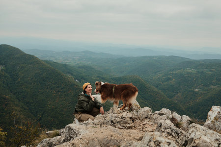 A smiling woman kneels on rocky cliffs as her dog gives her a paw with mountain landscapes in the background. Hiking with pets conceptの写真素材