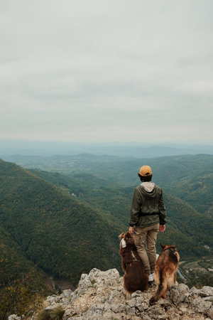 A woman with her two dogs Australian and German Shepherd stands on the rocky cliffs of Mount Kablar, Serbia, surrounded by green mountains and misty valleys. Hiking with pets concept.の写真素材