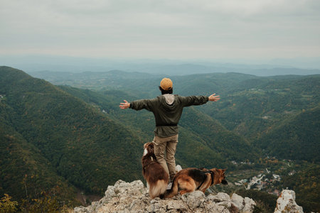A happy woman raises her arms on the top of Mount Kablar, Serbia, with two dogs Australian and German Shepherd beside her, enjoying the freedom of the mountains. Rear view. Hiking with pets conceptの写真素材