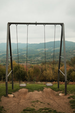 Two empty swings overlooking green mountains and autumn forest under cloudy sky. Perfect for travel, nostalgia, and peaceful outdoor visualsの写真素材