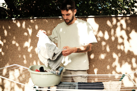 Young man standing outdoors sorting clothes from a plastic basin before hanging them on a drying rack. Concept of everyday home chores and responsibilityの写真素材