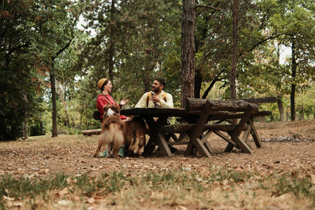 Young couple sitting and talking at a wooden picnic table in the forest with their dogs nearby....