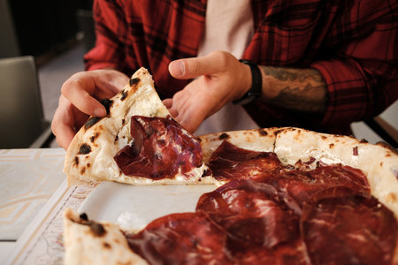 Close up of a man in a red checkered shirt enjoying pizza with cured meat at an outdoor cafeの写真素材