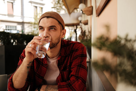 Young man in red checkered shirt and beanie drinking water at a cafe terraceの写真素材