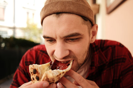 Young man biting into slice of pizza while sitting at cafe table in city centerの写真素材