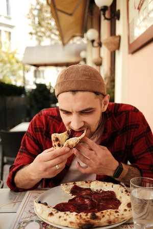 Young man biting into slice of pizza while sitting at cafe table in city centerの写真素材