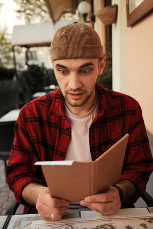 Surprised man in red checkered shirt reading restaurant menu at outdoor cafe table
