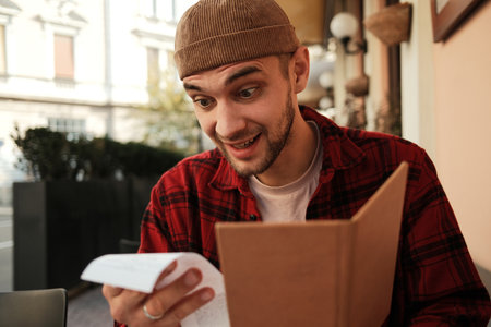 Amused young man looking at restaurant bill and laughing at outdoor cafe tableの写真素材