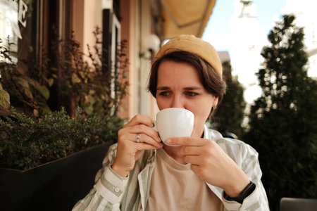 Woman in striped shirt and beige hat drinking coffee outdoors at sunny city cafe. Good morning in city conceptの写真素材