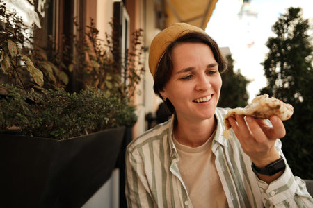 Happy woman smiling while holding pizza slice at outdoor cafe during sunny dayの写真素材