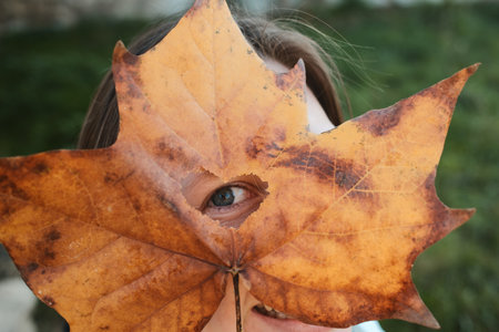Person playfully hides face behind large autumn leaf with heart-shaped hole showing one eye