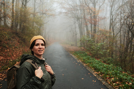 Smiling woman with backpack stands on quiet foggy road in autumn forest. Calm travel and exploration...