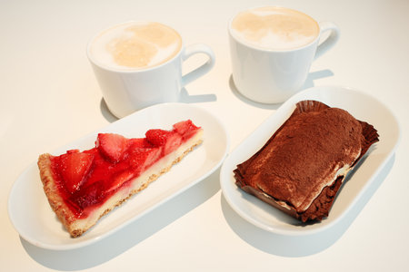 Two desserts, strawberry tart and tiramisu, paired with cups of cappuccino on a white minimalist table under warm light. The concept of breakfast in a cafeの写真素材
