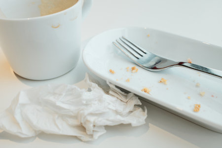 Empty cup with coffee foam, white plate with crumbs, fork, and crumpled napkin after a mealの写真素材