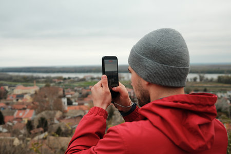 Man in grey beanie and red jacket recording city scenery with a smartphone. The frame reflects travel, technology, and personal experience. Travel blogger conceptの写真素材