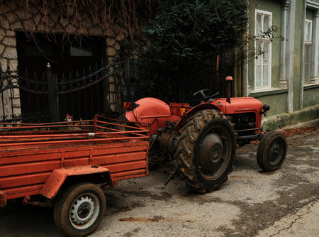 A red tractor with a small trailer stands on a quiet village road. The scene reflects rural work and agricultural lifestyleの写真素材