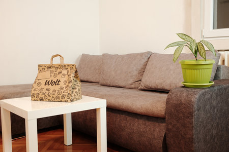 A Wolt paper delivery bag stands on a white table in a cozy living room with a grey sofa and a green plant. Modern food delivery lifestyle concept. Belgrade, Serbia - 22 November 2025のeditorial素材