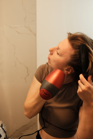A close-up of a woman drying her hair with a red hairdryer, concentrating on styling during a calm morning routineの写真素材