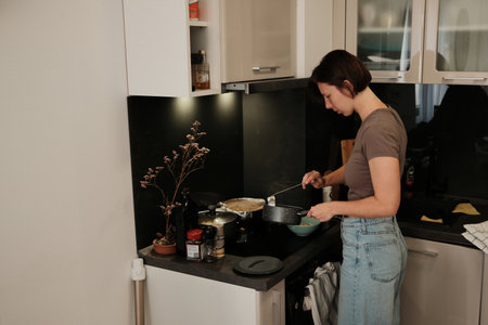 A woman prepares food in a modern kitchen, carefully ladling hot soup into a bowl as part of her daily cooking routineの写真素材