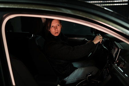 A woman drives a car out of a dim underground garage, illuminated by soft artificial light. The scene reflects daily commuting, independence, and modern mobility.の写真素材