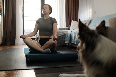 A woman meditates in a cross-leg position on a mat while a border collie observes from the side. The setting reflects calm energy, focus, and peaceful indoor routineの写真素材