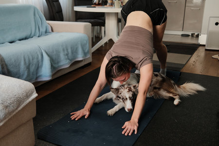 A woman performs a downward dog pose while a border collie lies directly under her. The moment captures humor, focus, and home wellnessの写真素材