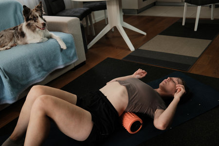 A woman lies on a foam roller while the border collie watches her from the sofa. The atmosphere shows home wellness with a peaceful pet presenceの写真素材