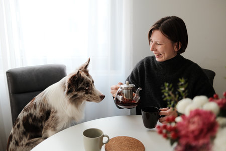 A woman laughs while holding a teapot as her dog curiously watches, capturing a warm and playful domestic interaction. The scene shows companionship and relaxed morning moodの写真素材
