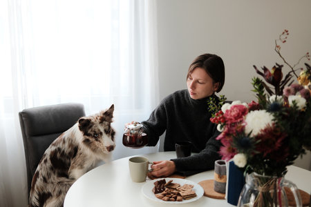 A woman pours hot tea into a cup while her dog sits beside her at the table. Concept of cozy domestic morning routineの写真素材
